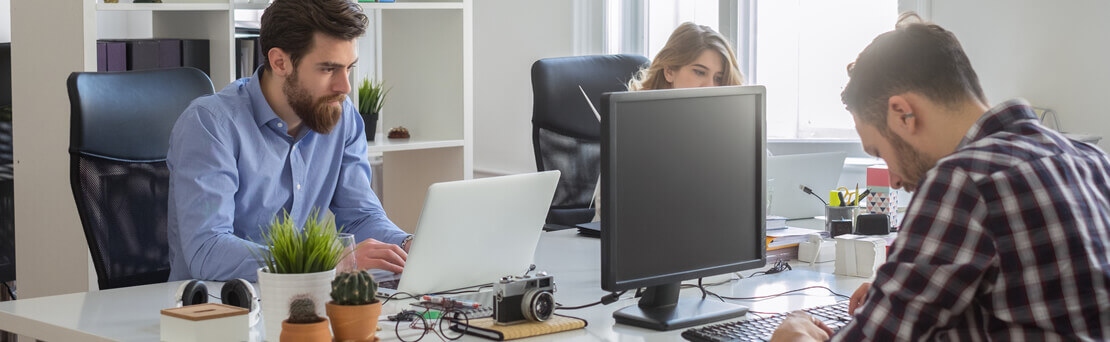 Two men and one woman working on computers at shared desk workspace.